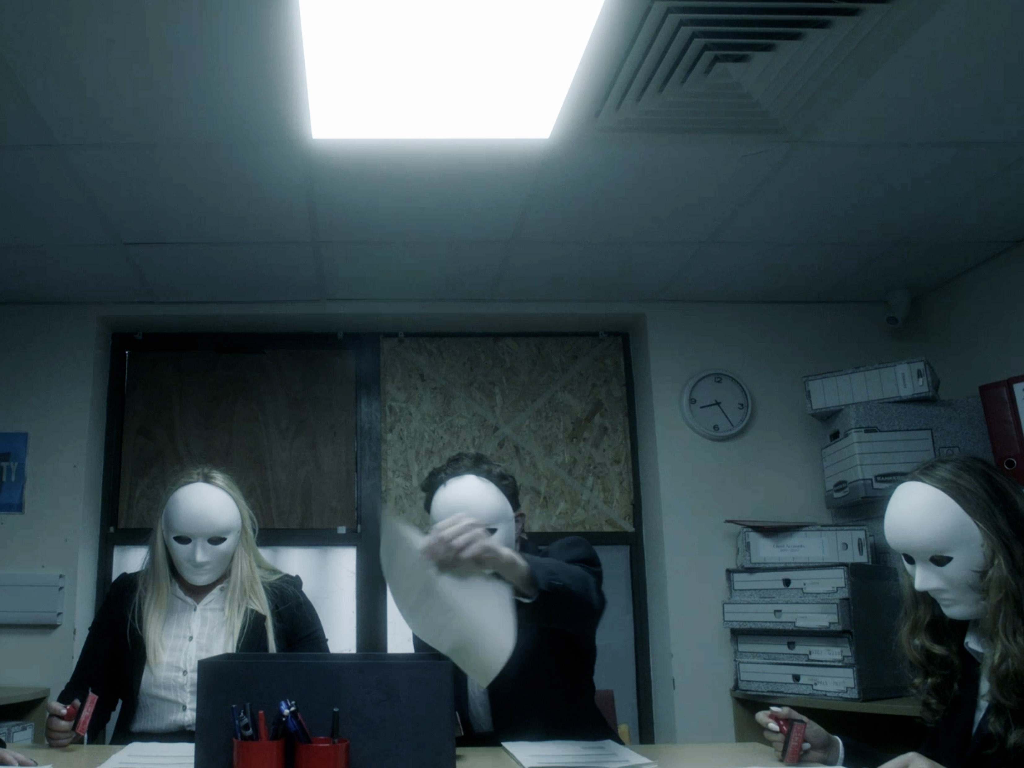 Production still from Typical? showing three masked figures seated at an office table under harsh overhead lighting.